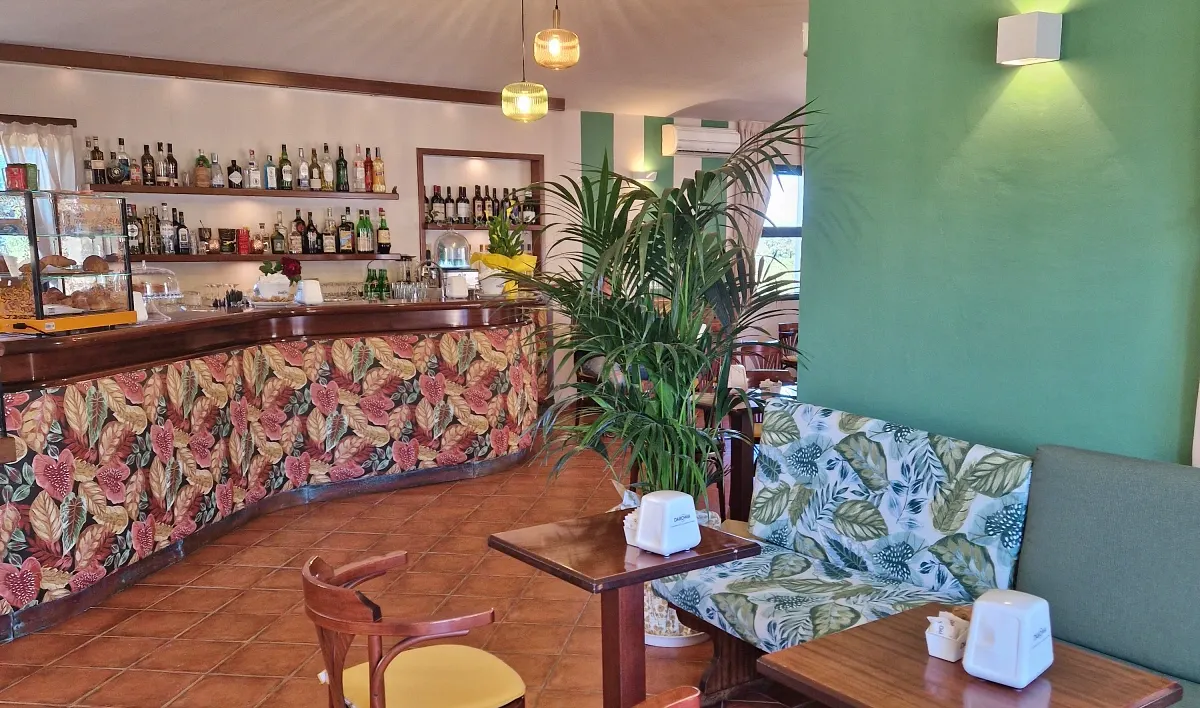 Bancone bar con rivestimento a foglie, scaffali di bottiglie e vetrina dolci; tavolini e divanetto verde